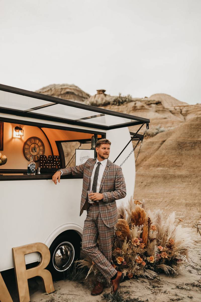 Man in brown plaid suit leaning against white mobile bar with drink in hand