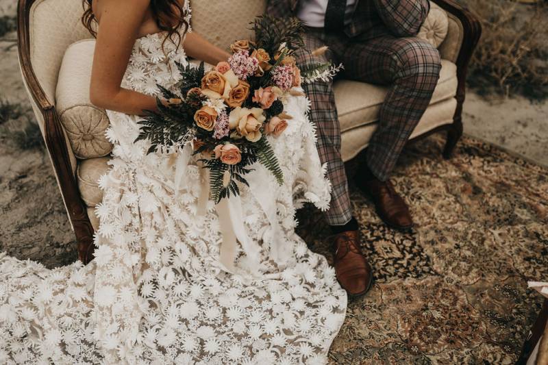 Bride and groom sit on couch with large white lace dress strewn holding bouquet 