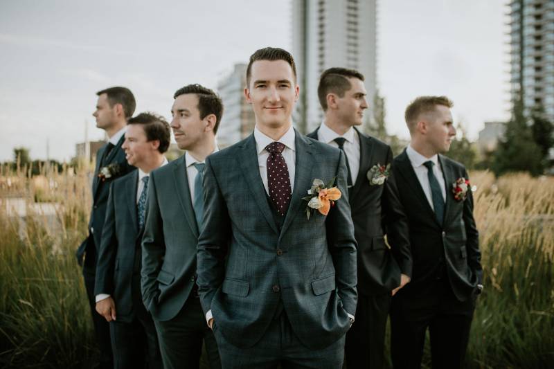 Groom and groomsmen stand to side looking over shoulder in wheat field 