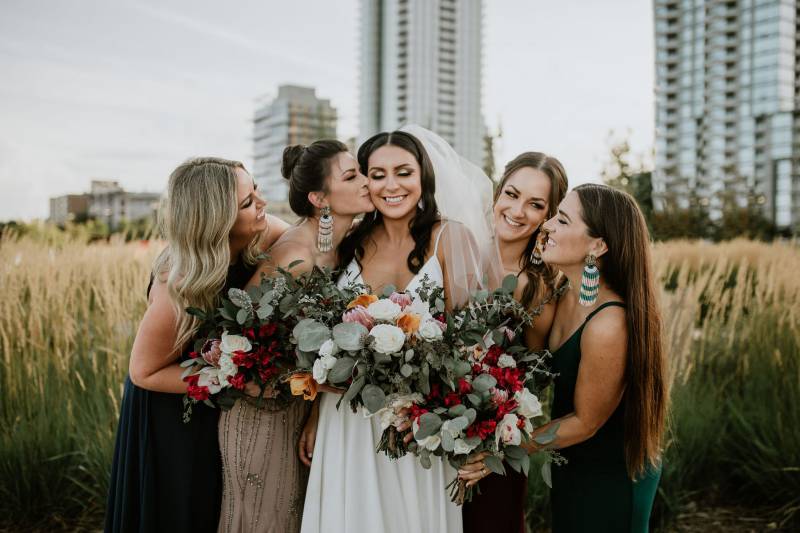 Bride and bridesmaids smile together holding white and red bouquets in grassy field 