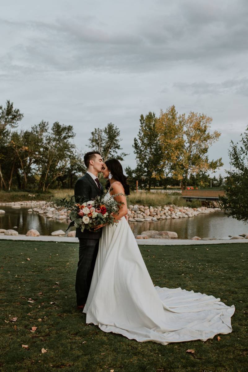 Groom kisses brides forehead holding bouquet in grassy field by pond