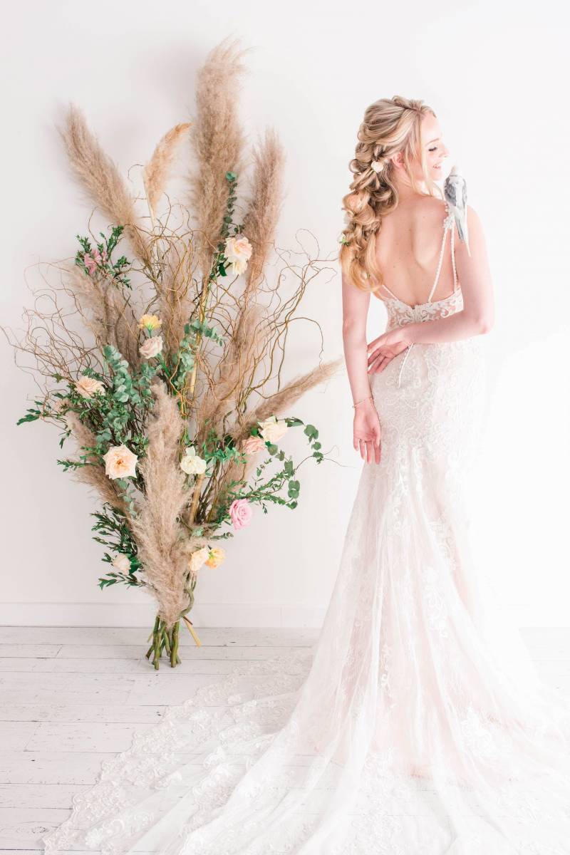 Bride in white lace openback dress with white cockatoo on shoulder beside wheatgrass floral arrangement on wall 