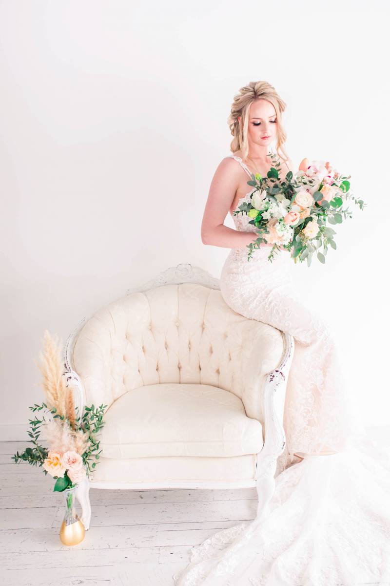 Bride holding blush white and pink bouquet sitting on edge of white seat and gold vase 