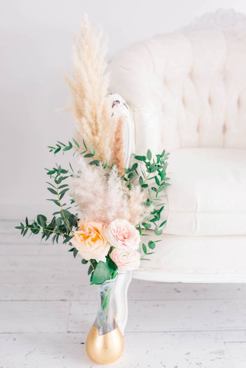 Blush pink and yellow floral arrangement with papas grass accent in gold bottom vase in front of white couch 