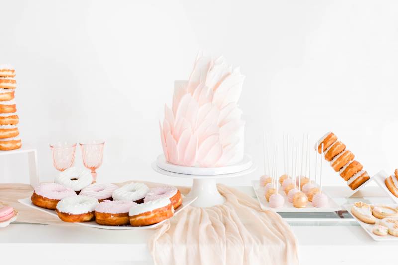 Pale pink floral wedding cake beside pin k and white baked goods on white table