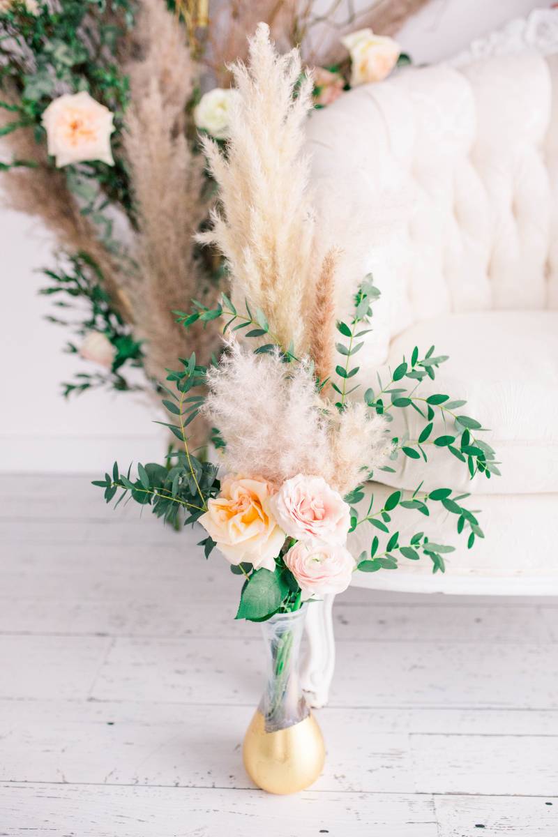 Blush pink and yellow floral arrangement with papas grass accent in gold bottom vase in front of white couch 