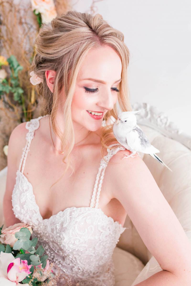 Bride in white lace dress smiling looking at cockatoo on shoulder 