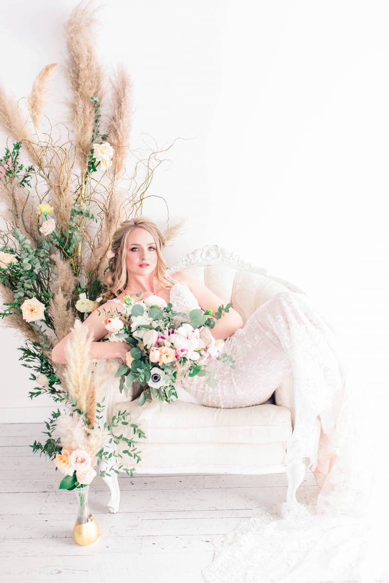 Bride holding blush white and pink bouquet sitting on white seat beside pampas grass arrangement 