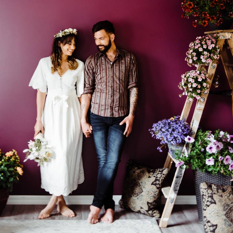 Man and woman lean against burgundy wall smiling holding hands beside ladder with flowers 