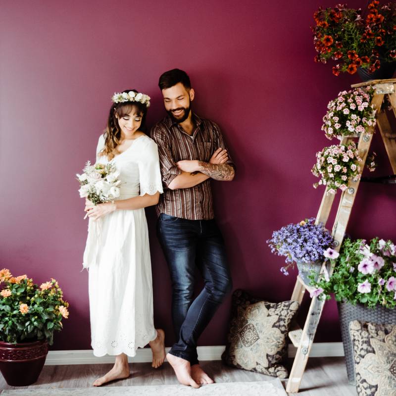 Man and woman smile leaning against burgundy wall beside ladder and flowers  