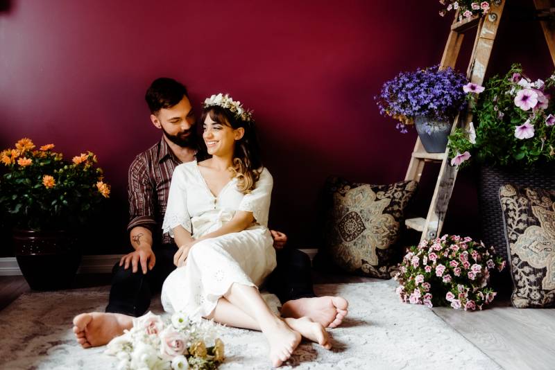Woman in white flower crown sits in mans lap barefoot beside wooden ladder with flowers 
