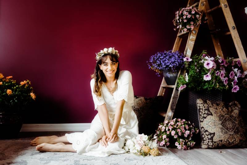 Woman sits in white dress and white flower crown beside wooden ladder with flowers 