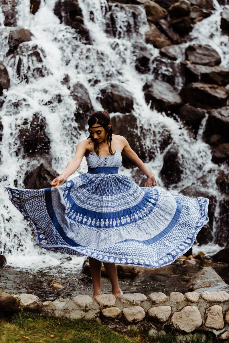 Woman spins blue and white dress in front of rocky waterfall 