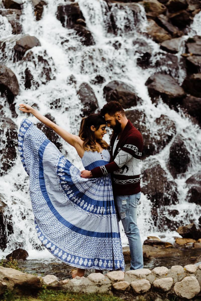 Woman throws blue skirt behind while man embraces in  front of rocky waterfall 
