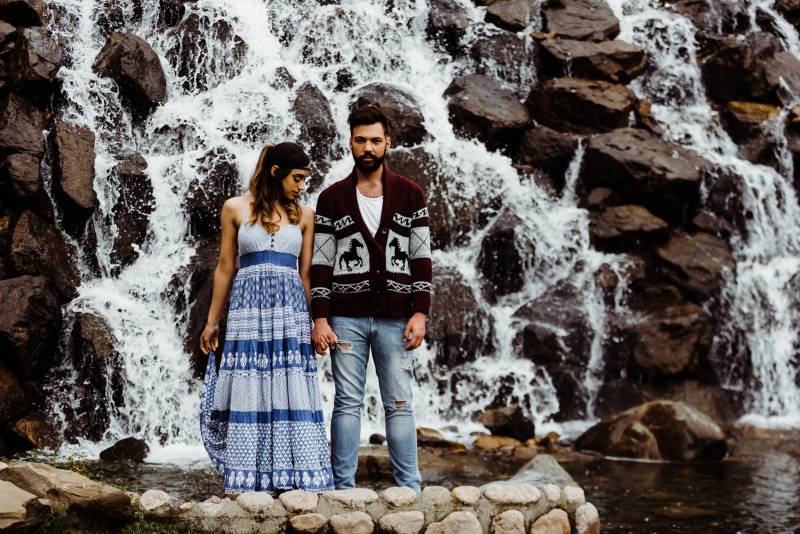 Man and woman stand holding hands in front of rocky waterfall 