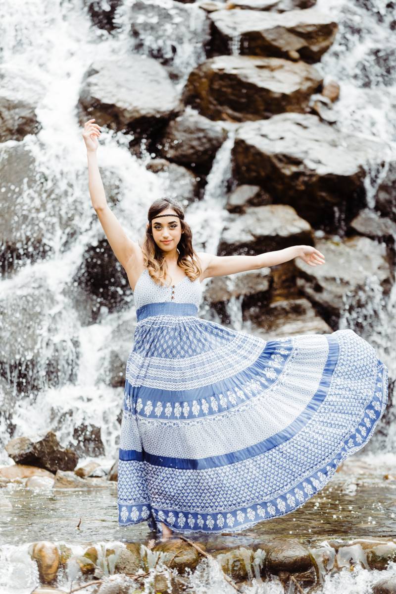 Woman throws blue skirt out with hands raised in front of rocky waterfall 
