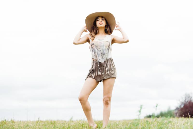 Woman stands knee cocked holding brim of large hat in grassy field 