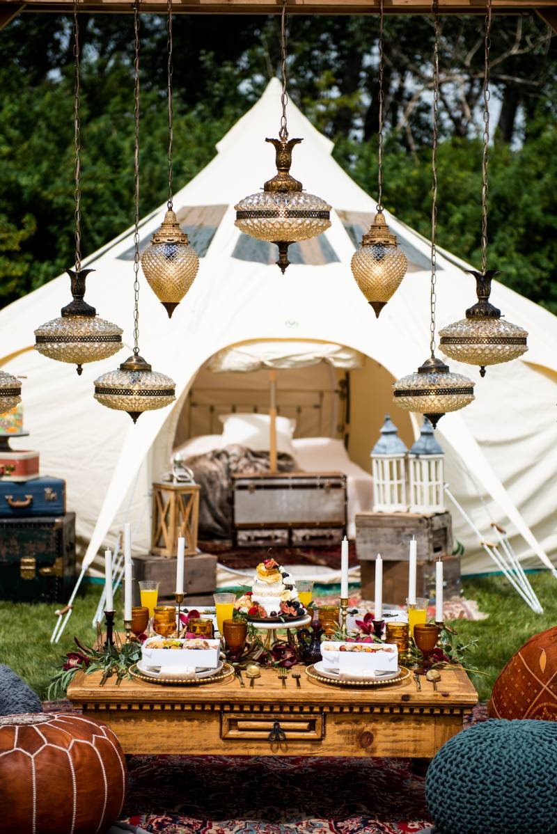 Boho pouf seating and small wooden table setting under small chandeliers in front of white tent