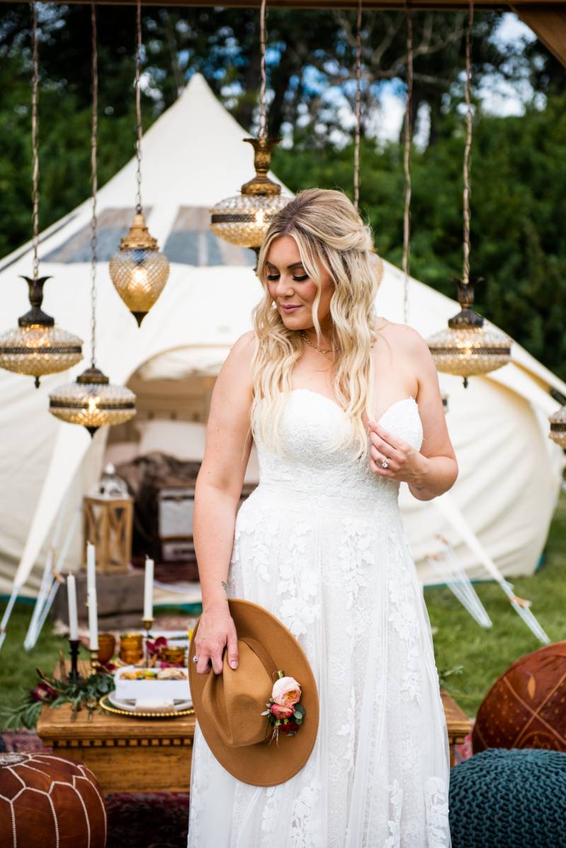 Woman stands in white dress holding brown hat in front of chandeliers and white tent 