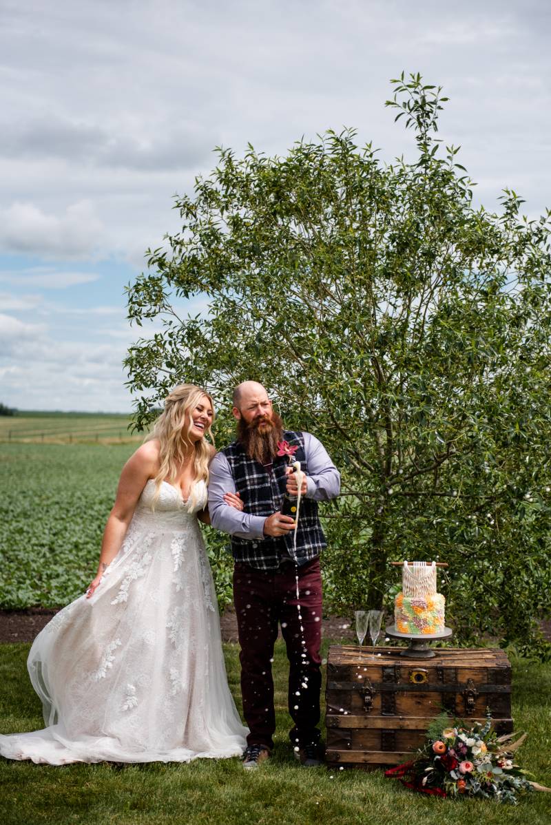 Man and woman laugh holding bottle of spilling champagne beside boho wedding cake 
