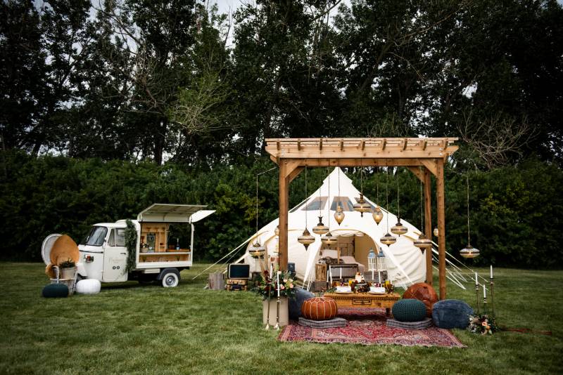Wooden wedding arch with hanging chandeliers and poufs in front of white tent and food truck  