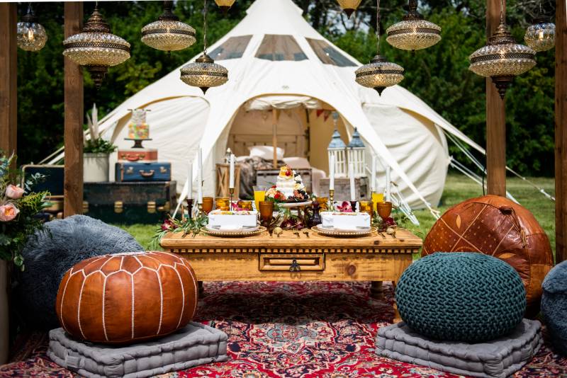 Boho pouf seating and small wooden table setting under small chandeliers in front of white tent