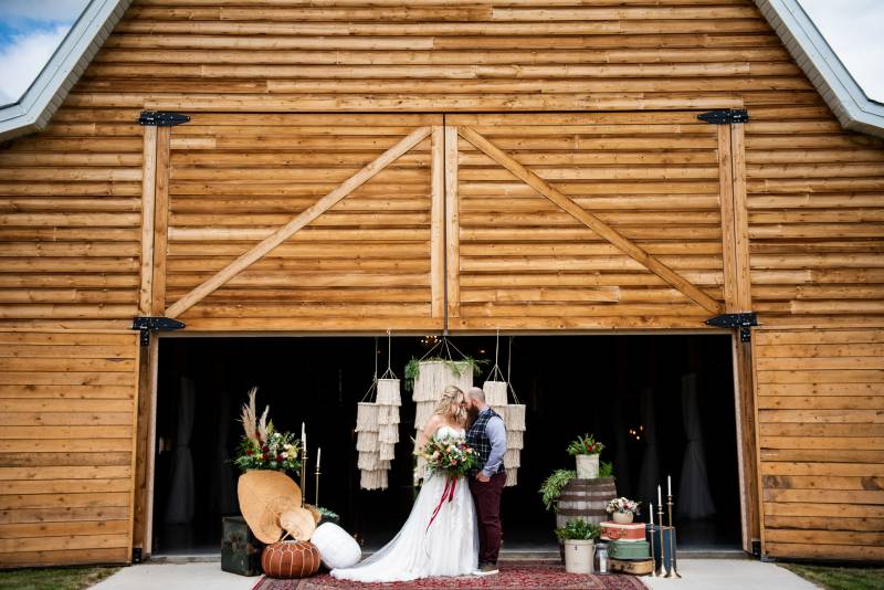 Man and woman hold hands in front of large wooden barn and hanging macrame chandeliers 