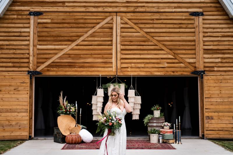 Woman in white dress holding orange and maroon bouquet standing in front of wood barn 