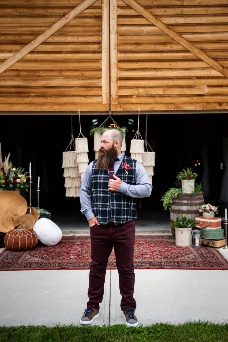 Man standing with plaid vest and maroon pants in front of wooden barn doorway