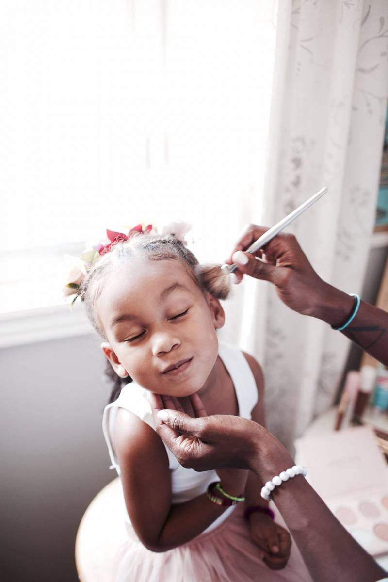 Child in blush dress closes eyes while having makeup applied