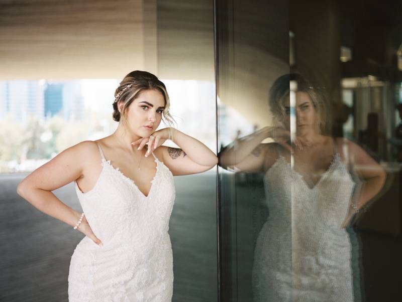 Bride leaning against glass wall hand on hip wearing white lace dress  