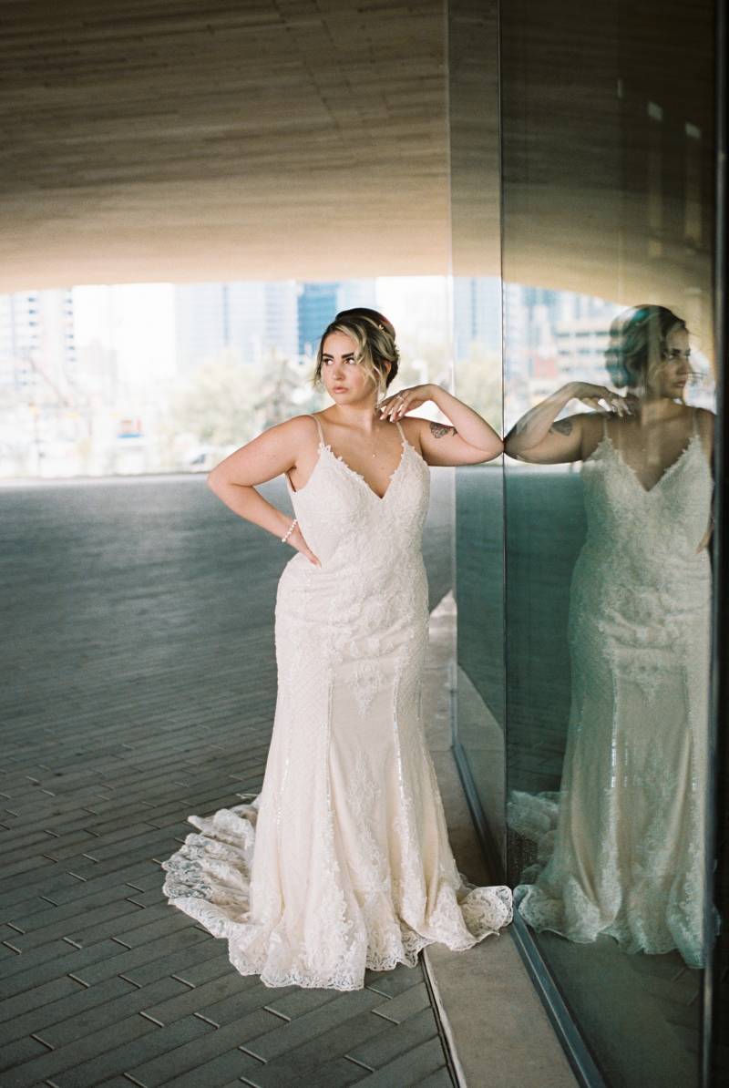 Bride leaning against glass wall hand on hip wearing white lace dress  