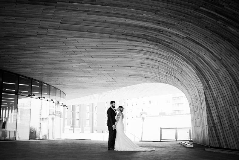 Bride and groom stand holding hands under archway 