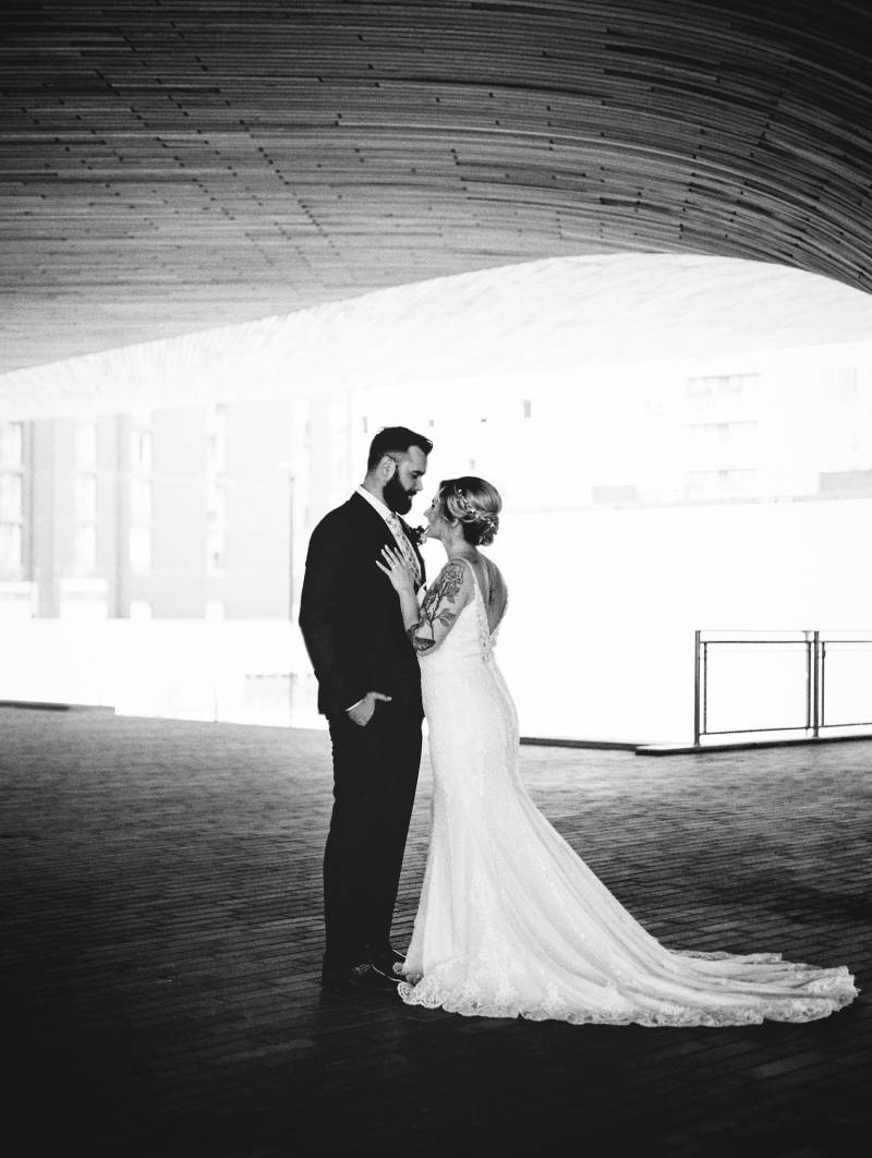 Bride leans against groom chest while hands in pockets black and white 