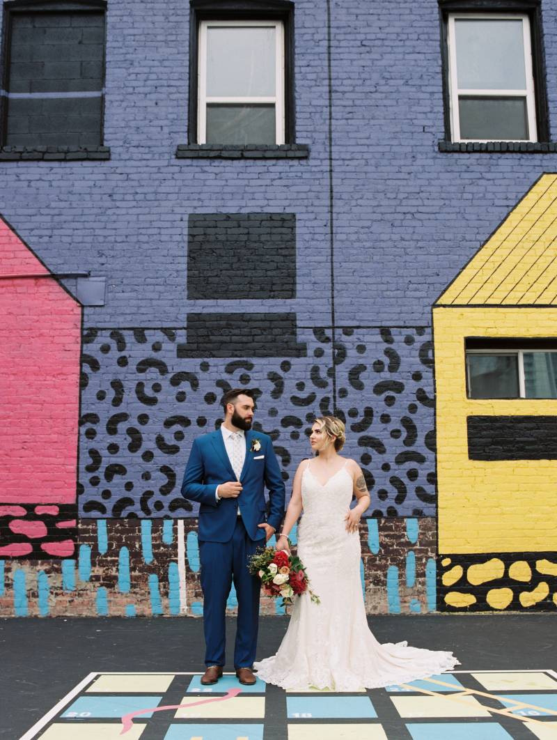 Bride and groom stand looking at each other against painted brick building 