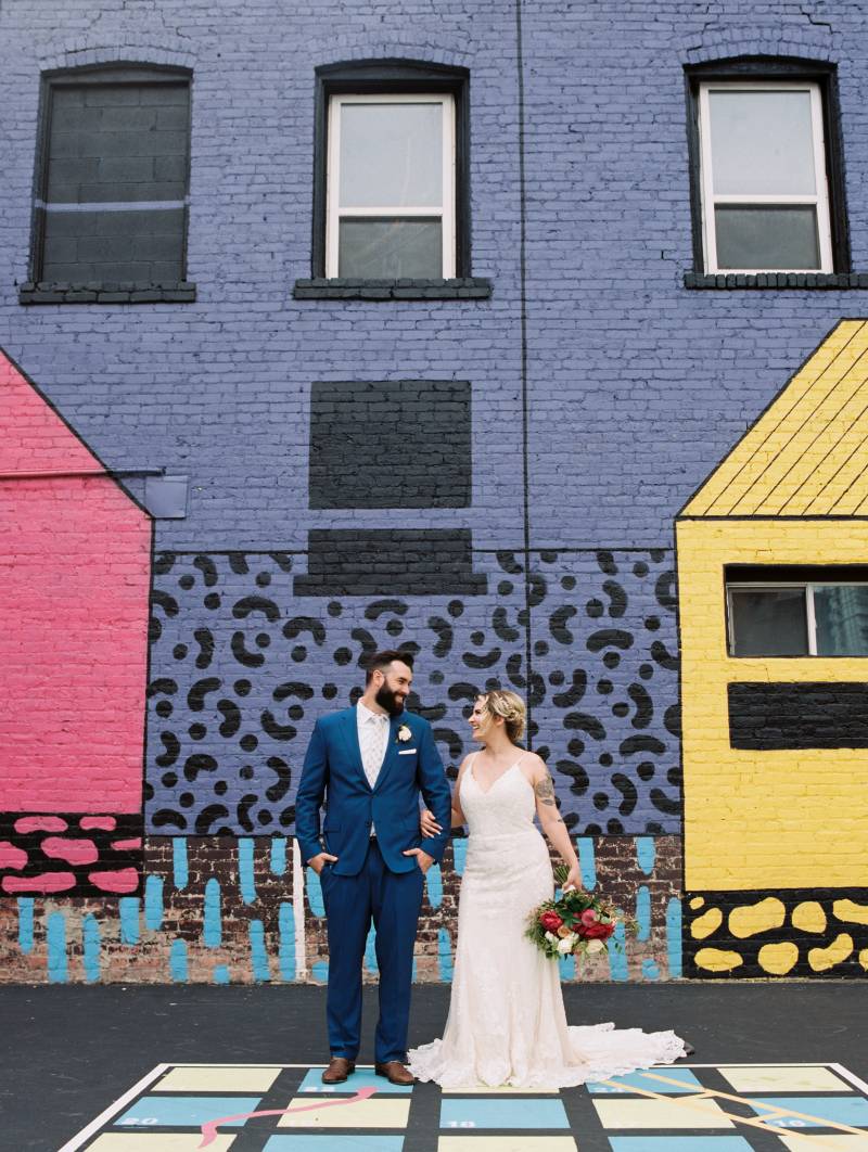 Bride and groom smile looking at each other holding bouquet against painted brick building  
