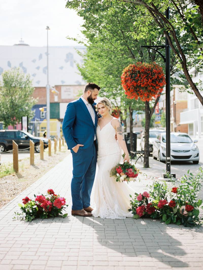 Bride and groom stand together on sidewalk beside red flowers and hanging potted flowers 