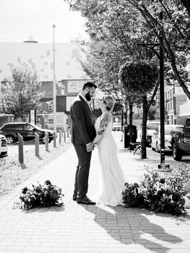 Bride and groom stand together on sidewalk beside red flowers and hanging potted flowers 