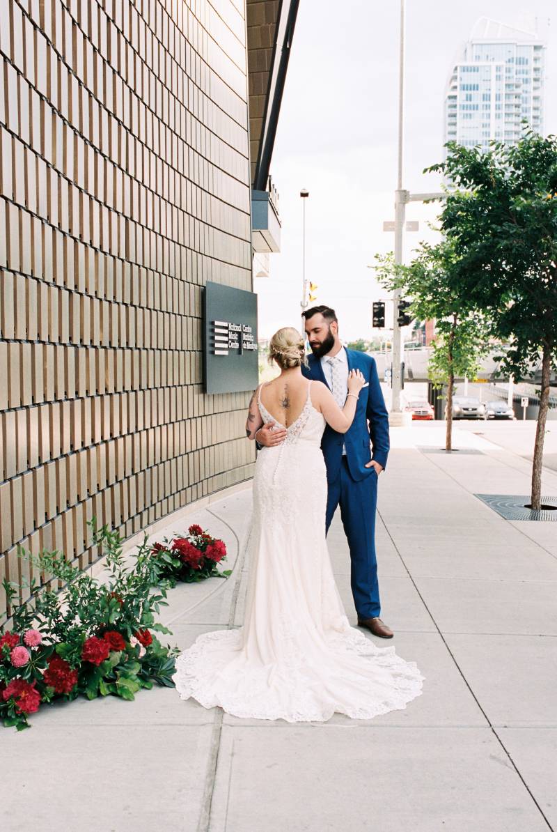 Bride and groom embrace beside building and red floral arrangement to side 