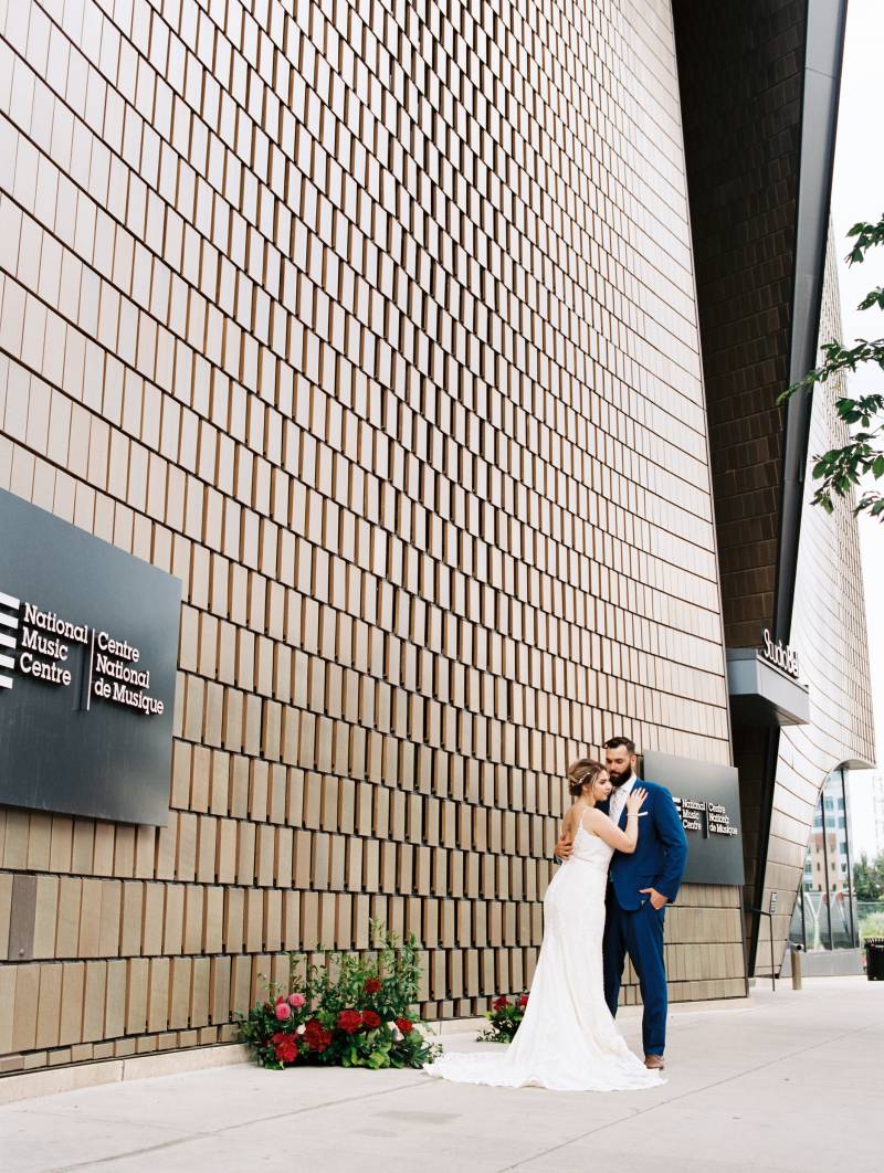 Bride and groom embrace leaning against chest beside building and red floral arrangement to side 