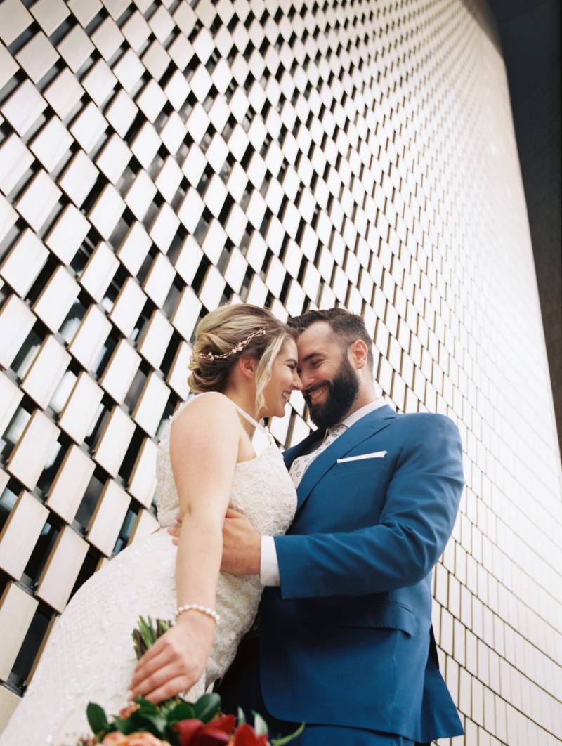 Bride and groom embrace smiling holding bouquet beside slated wall 