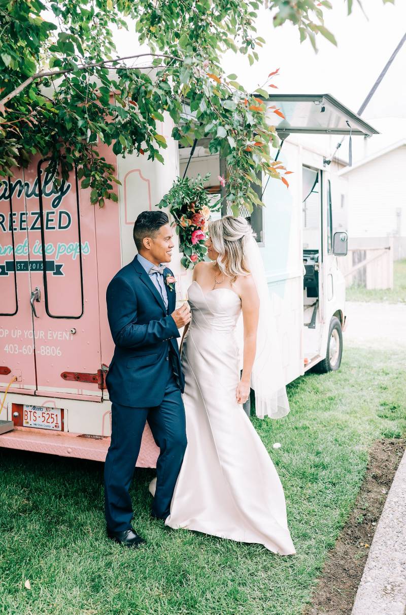 Bride and groom stand facing leaning against white and pink food truck 