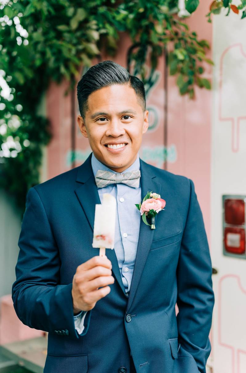 Groom stands in dark blue suit holding ice pop smiling