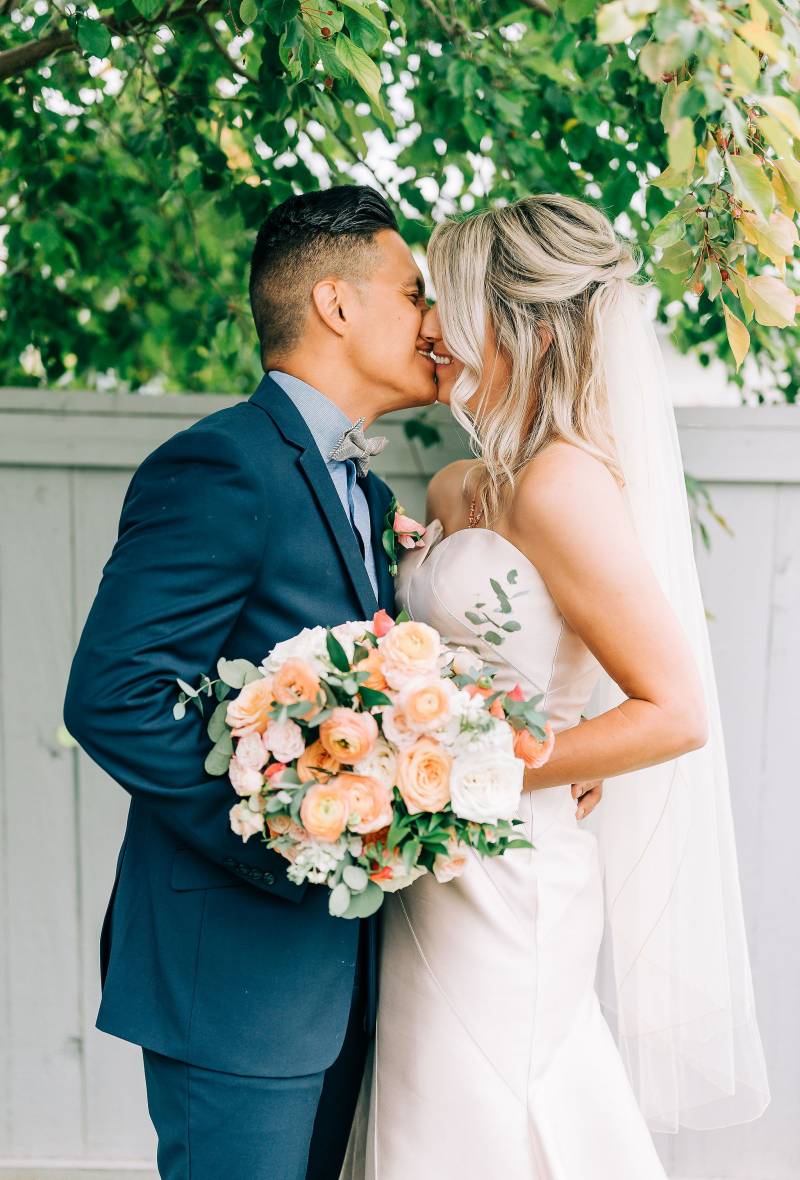 Bride and groom smile while kissing holding peach and white bouquet 