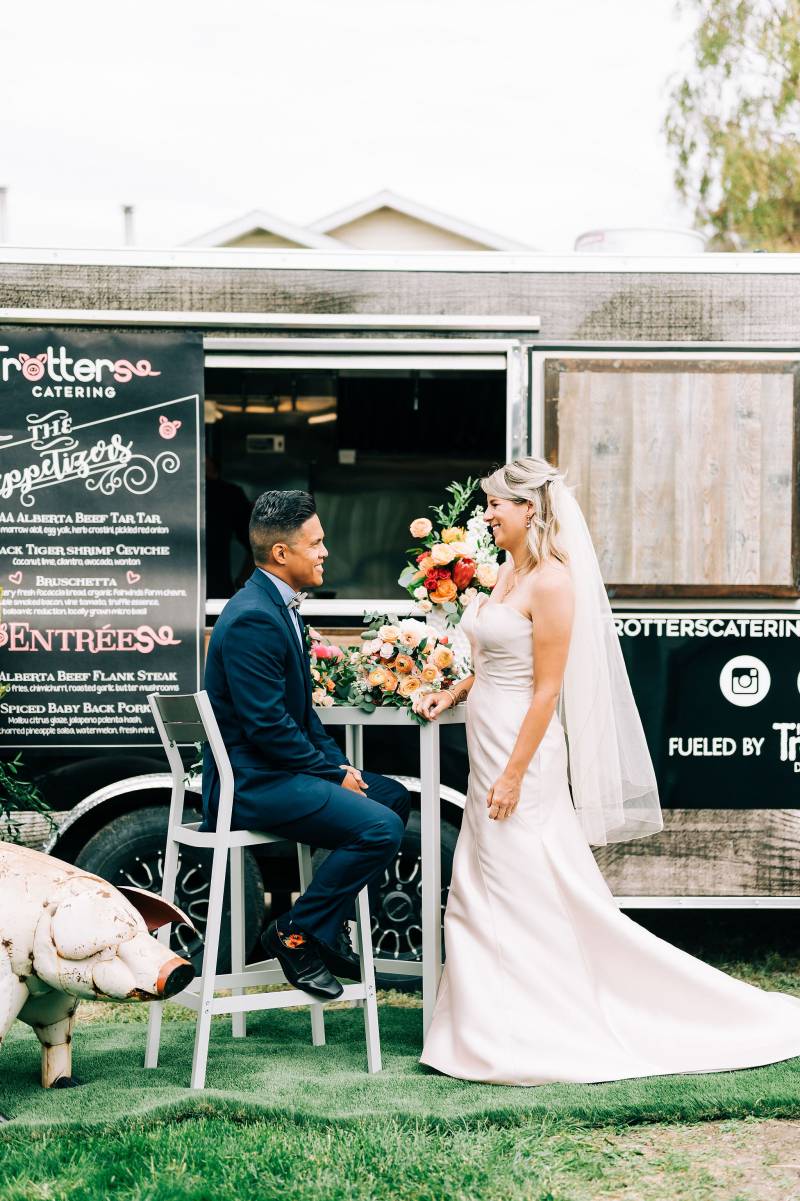 Bride in long white dress and veil standing facing groom sitting in front of food truck 