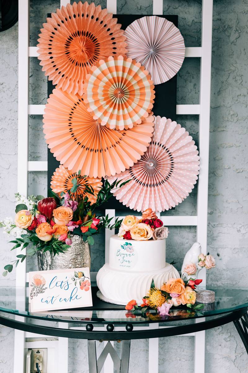 Orange and peach paper fans on latus over glass table with white wedding cake with floral accents  