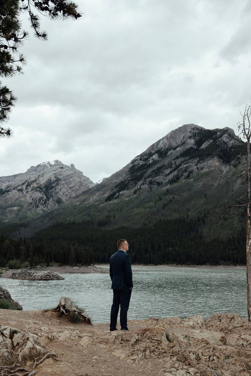 Groom standing with hands in pockets overlooking lake and mountains 