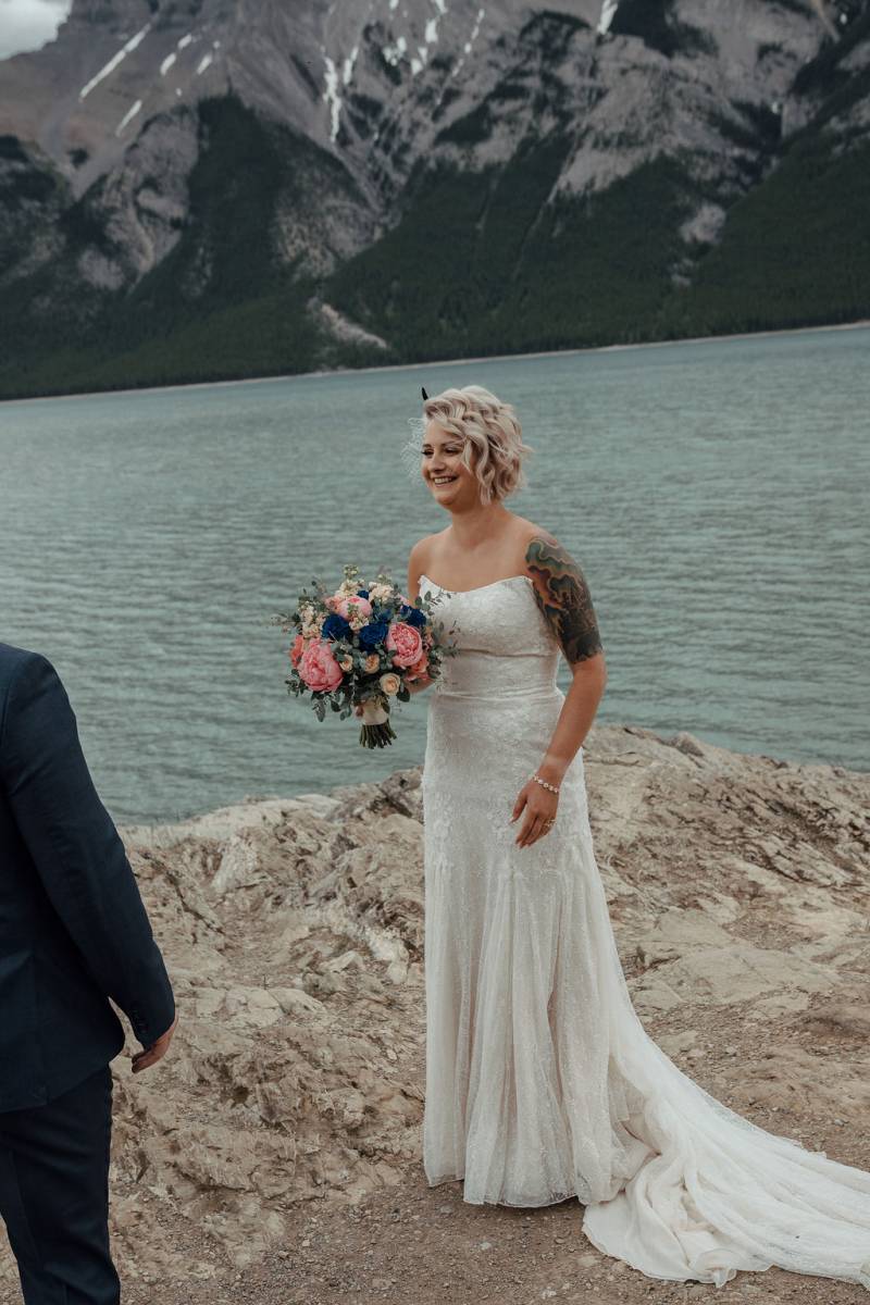 Bride holding bouquet smiling standing on rock overlooking lake and mountain 