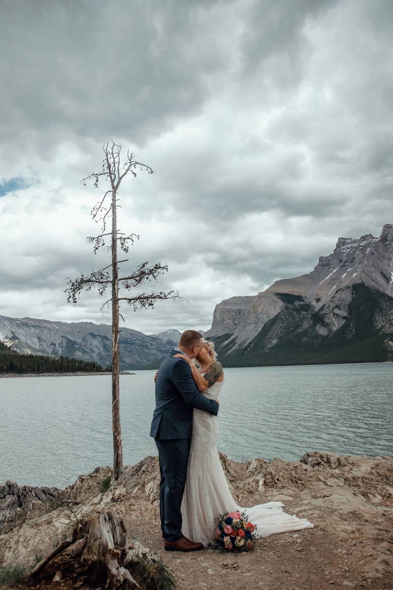 Bride and groom kiss while embracing on rocky surface overlooking mountains and lake 