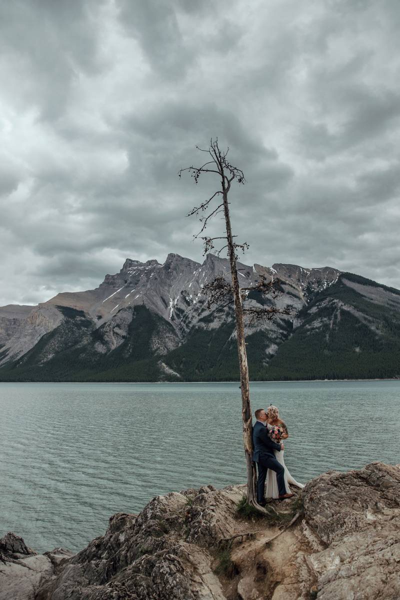 Bride and groom kiss leaning on dead tree overlooking lake and mountains 